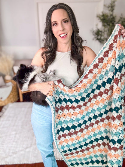 Smiling woman holding a small fluffy dog and a rust, peach and teal crochet granny stitch blanket