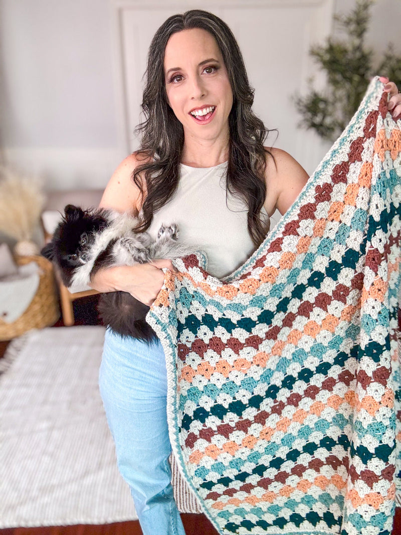 Smiling woman holding a small fluffy dog and a rust, peach and teal crochet granny stitch blanket