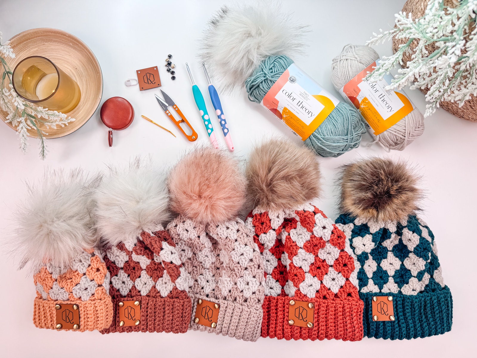 A row of five crocheted pom-pom hats in various colors on a white surface, alongside yarn, crochet tools, labels, and a beverage in a mug—an ideal setting for crafting handmade gifts.