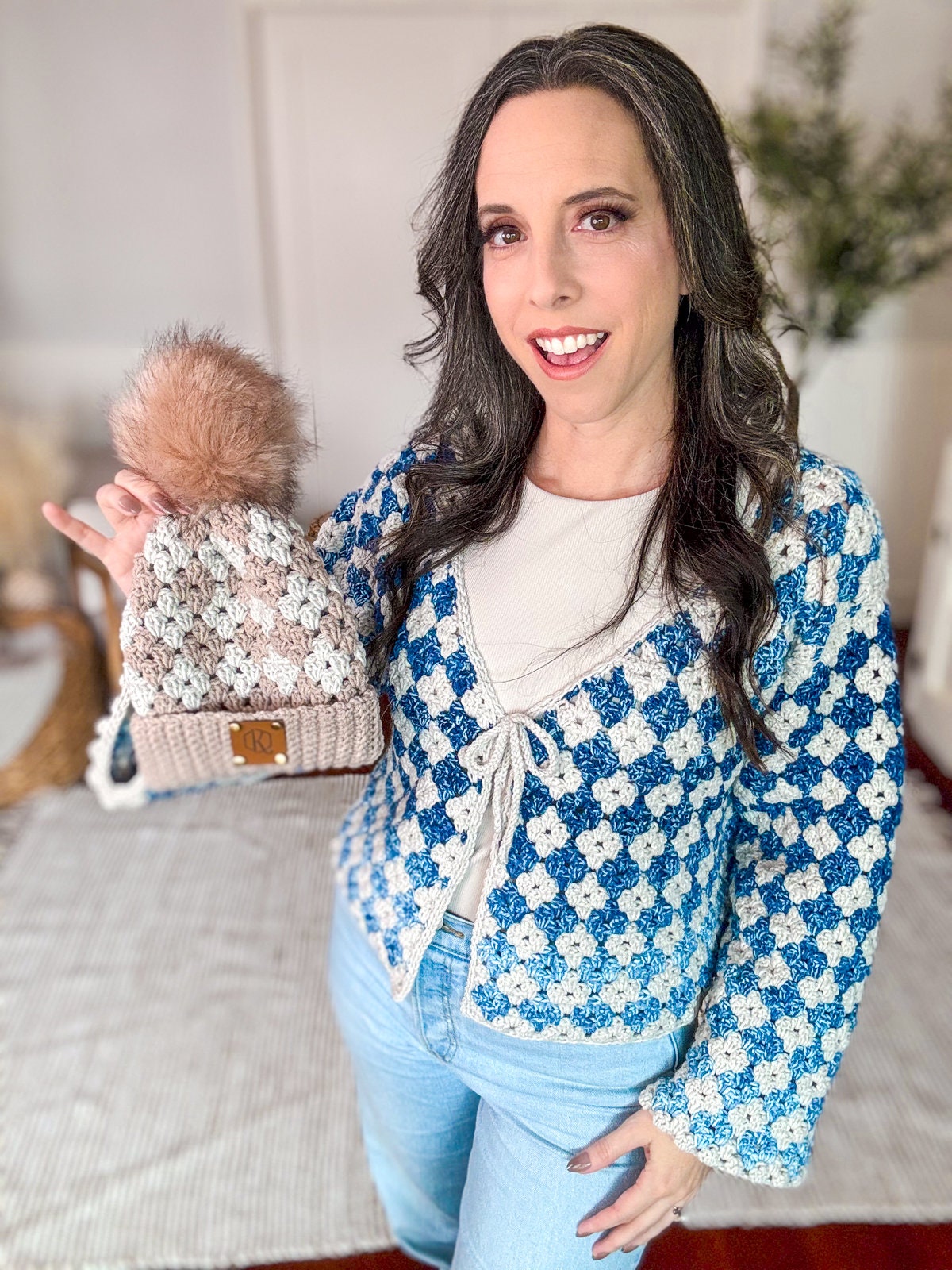 Person with long hair wearing a blue and white checkered cardigan and pale blue jeans, holding a crocheted hat with a large pom-pom. They exude charm with their unique jewellery and vintage clothes, making the hat feel like a handmade gift.