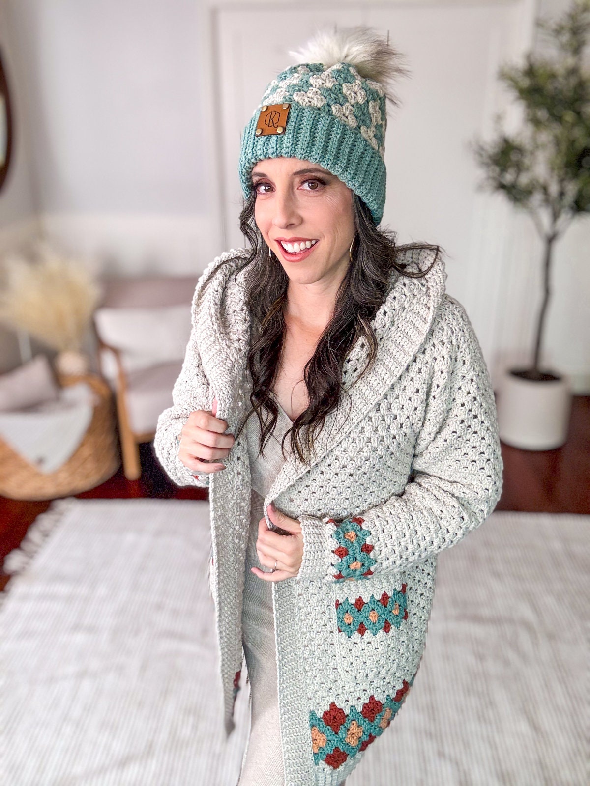 A woman stands indoors, wearing a crocheted sweater and matching beanie. She smiles while holding the sweater collar with one hand. The background features a chair and potted plant, adding to the charm of her vintage clothes ensemble.