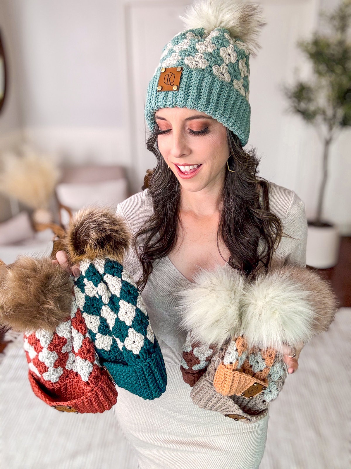 A woman wearing a knitted hat with a pom-pom holds three similar hats of different colors while smiling indoors, presenting them as the perfect handmade gift.