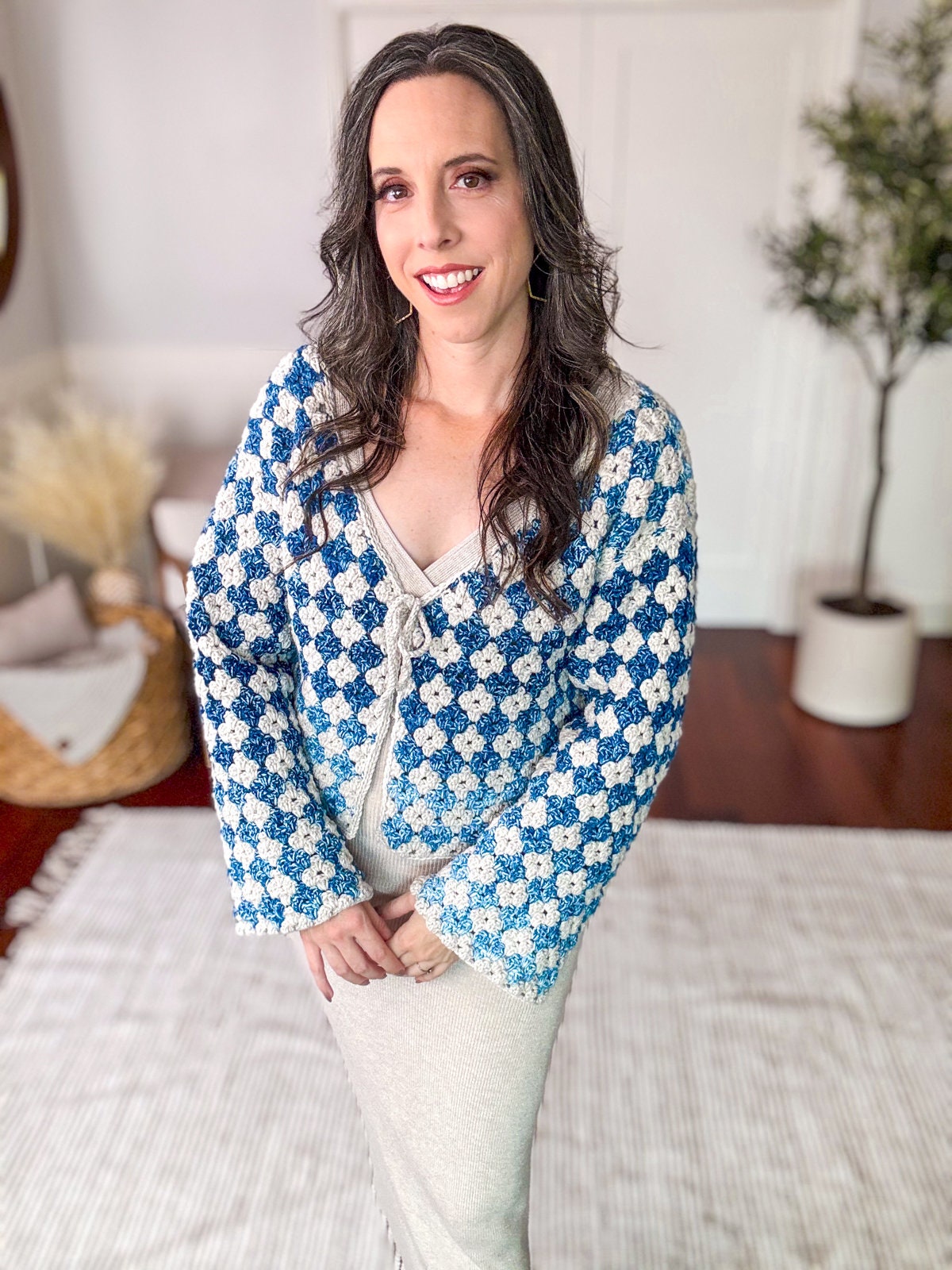 A woman with long dark hair stands indoors wearing a blue and white patterned cardigan and a beige dress, smiling at the camera. A plant and wicker basket are in the background, along with unique jewellery that adds a touch of charm.