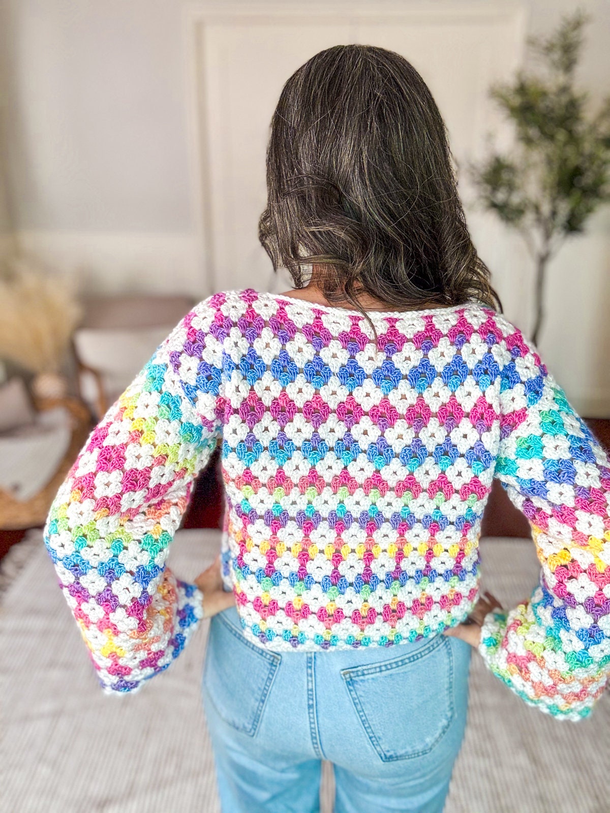 A person with long hair is standing indoors, facing away, wearing a colorful crocheted sweater with a multi-colored diamond pattern and light blue jeans. Their look is complemented by unique jewellery that accentuates the vintage clothes they wear.