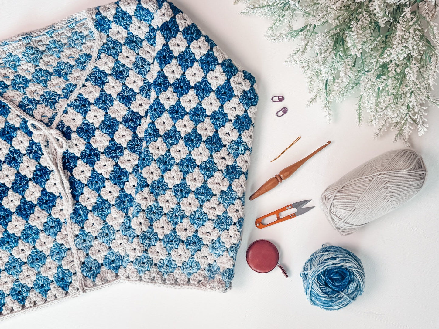 A crocheted blue and grey checkered blanket with crochet hooks, a tape measure, scissors, stitch markers, grey and blue yarn balls, and a snowy artificial plant on a white surface—a perfect handmade gift.