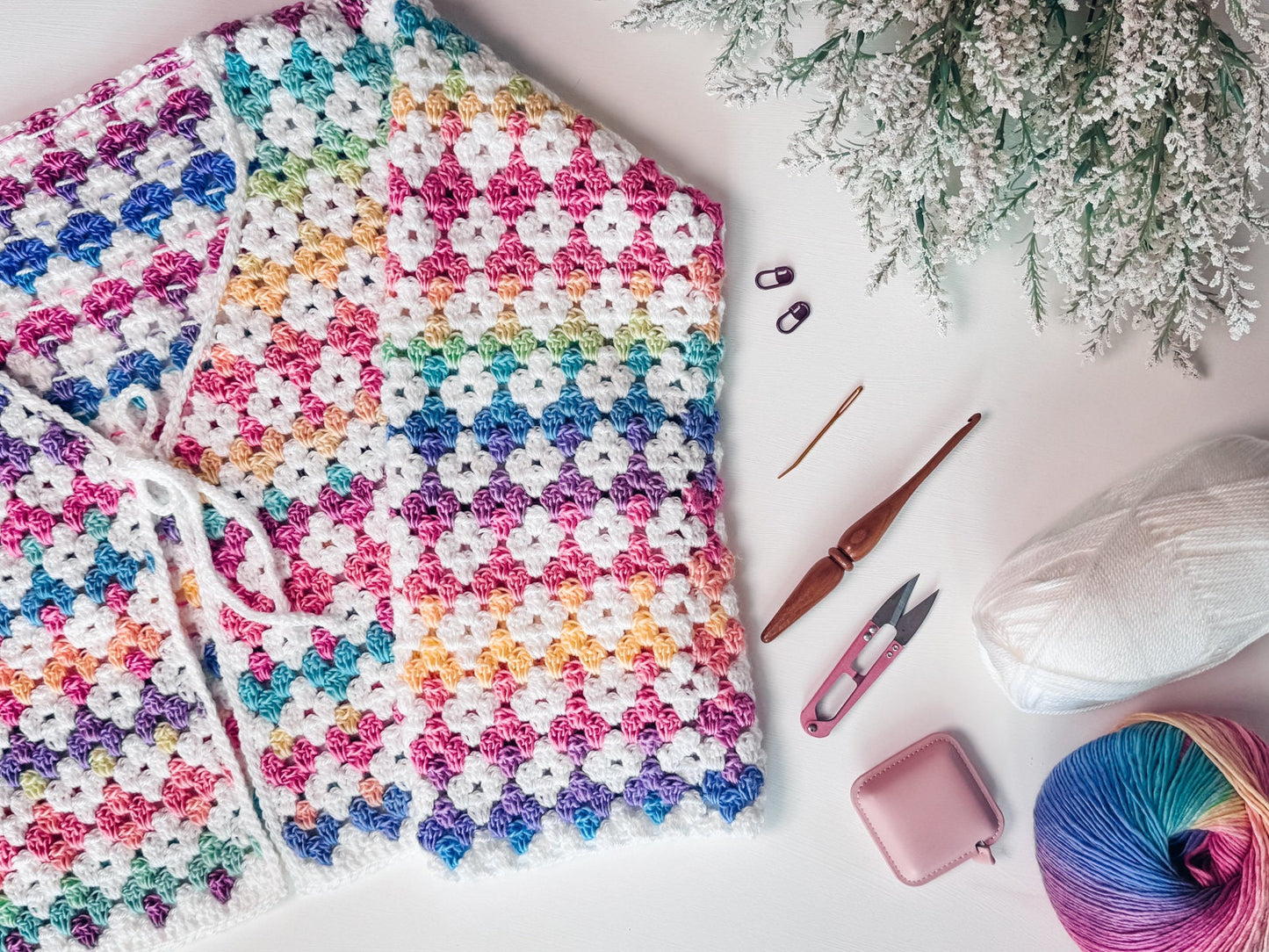 A colorful crocheted blanket, perfect as a handmade gift, is surrounded by crochet tools, vibrant yarn, and a plant on a white surface.