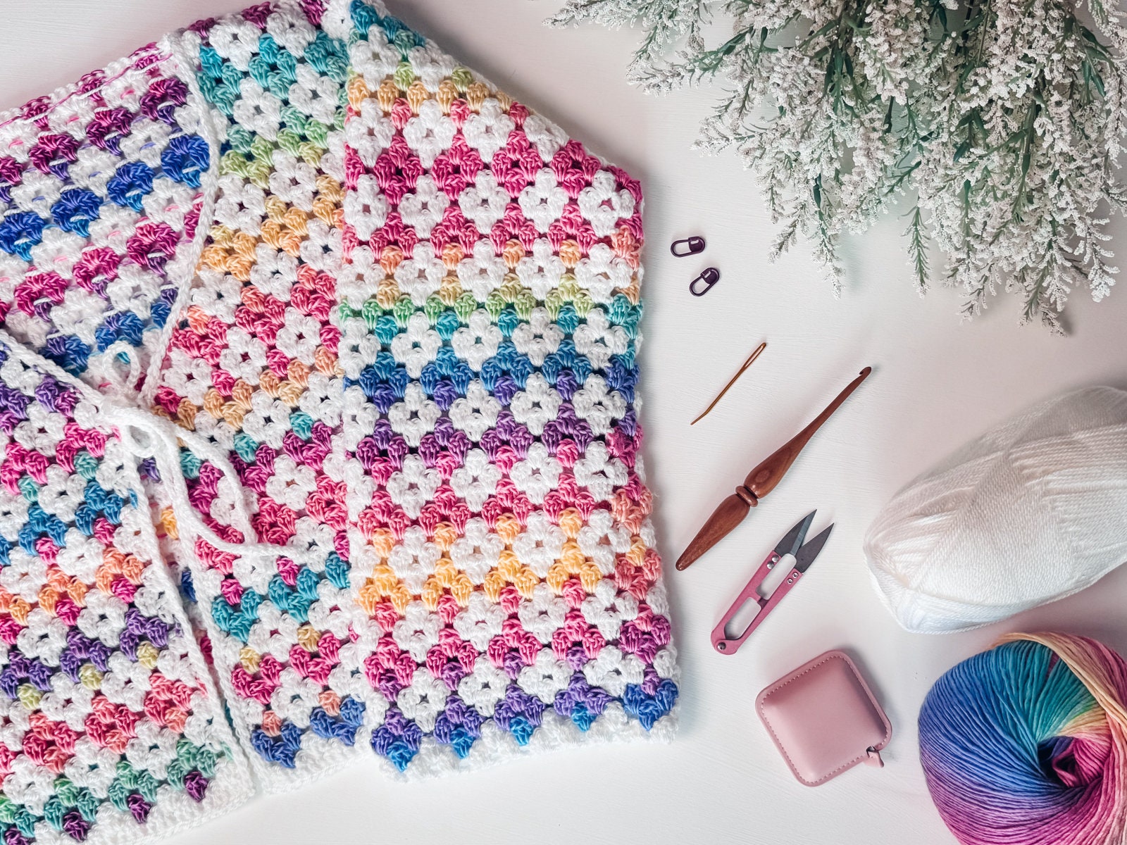 A colorful crocheted blanket, perfect as a handmade gift, is surrounded by crochet tools, vibrant yarn, and a plant on a white surface.