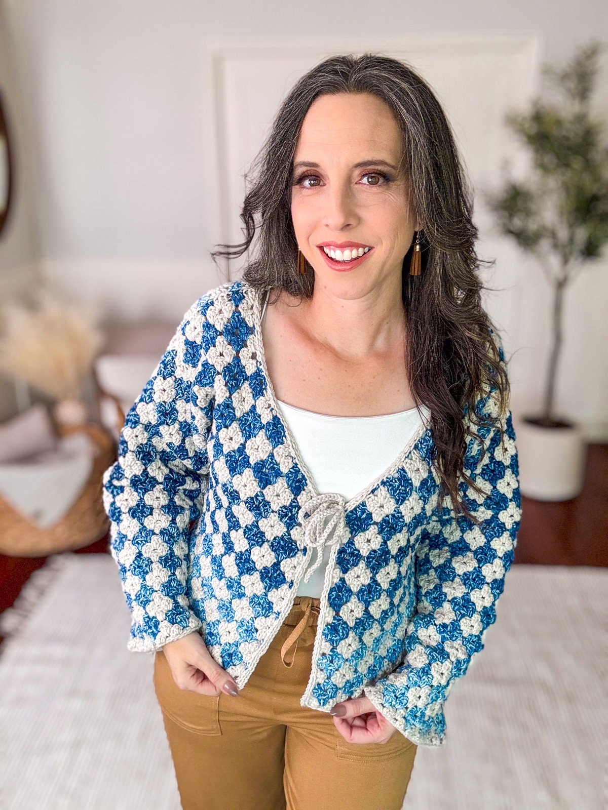 A person with long dark hair is smiling and wearing a blue and white checkered sweater over a white top, accessorized with unique jewellery, standing indoors.