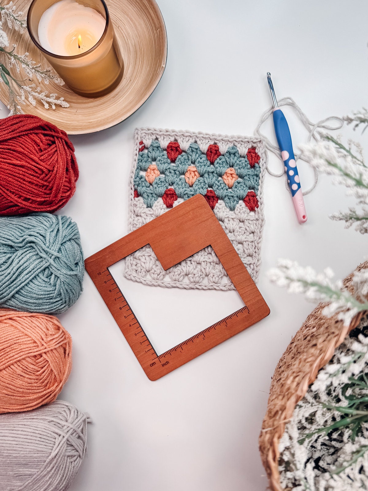 A crocheted square with a colorful pattern, balls of yarn in various colors, a crochet hook, and a square wooden ruler are arranged on a white surface. A lit candle is in the top left corner, making it the perfect setup for creating.