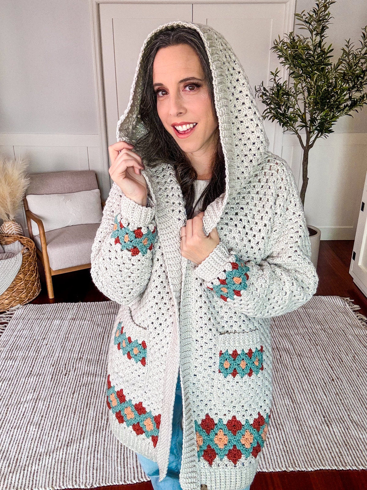 Person wearing a hooded, crocheted cardigan with colorful geometric patterns, standing indoors in a room with a beige armchair, a plant, unique jewellery and a textured rug.