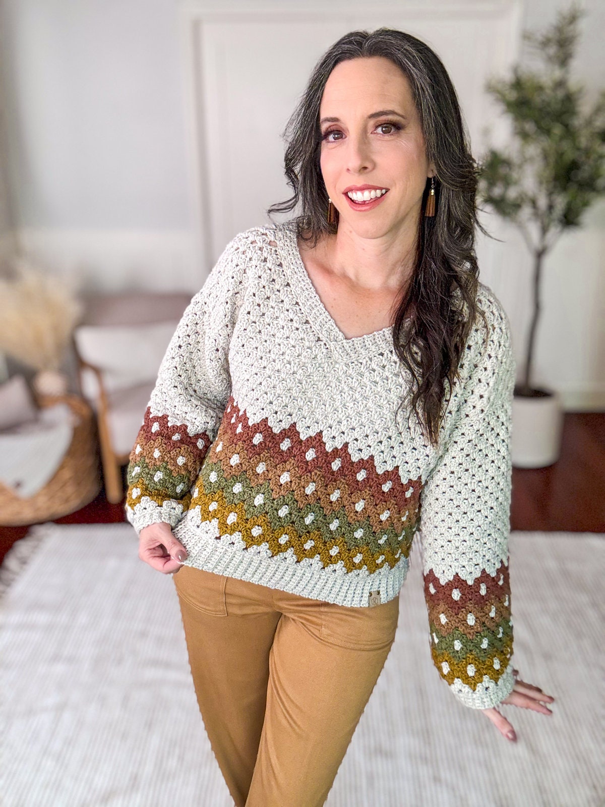 A woman in a crochet sweater with a green, brown, and white pattern and brown pants stands in a room with a plant and a basket in the background. She is smiling and looking at the camera, showcasing her unique jewelry that complements her perfectly.