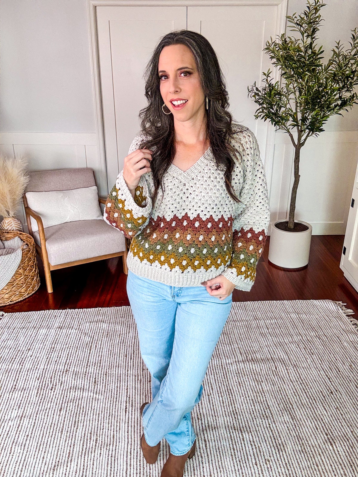 Woman standing indoors, wearing a knit sweater and light blue jeans, complemented by unique jewellery. A neutral-colored rug and indoor plants are visible in the background.