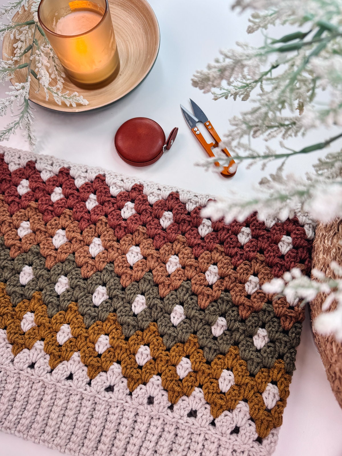 Crocheted multi-colored zigzag pattern swatch accompanied by scissors, a circular measuring tape, a lit candle, and greenery on a white surface—a perfect scene for creating unique jewellery or preparing a heartfelt handmade gift.