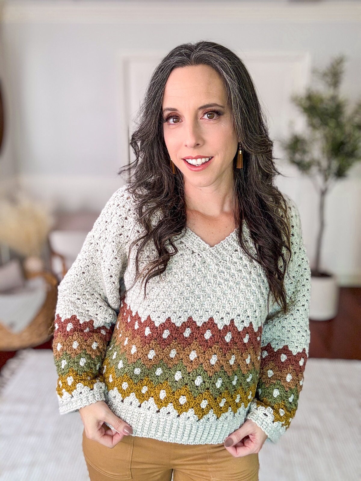 A woman wearing a multicolored knit sweater and beige pants stands indoors, smiling at the camera, showcasing her unique jewellery.