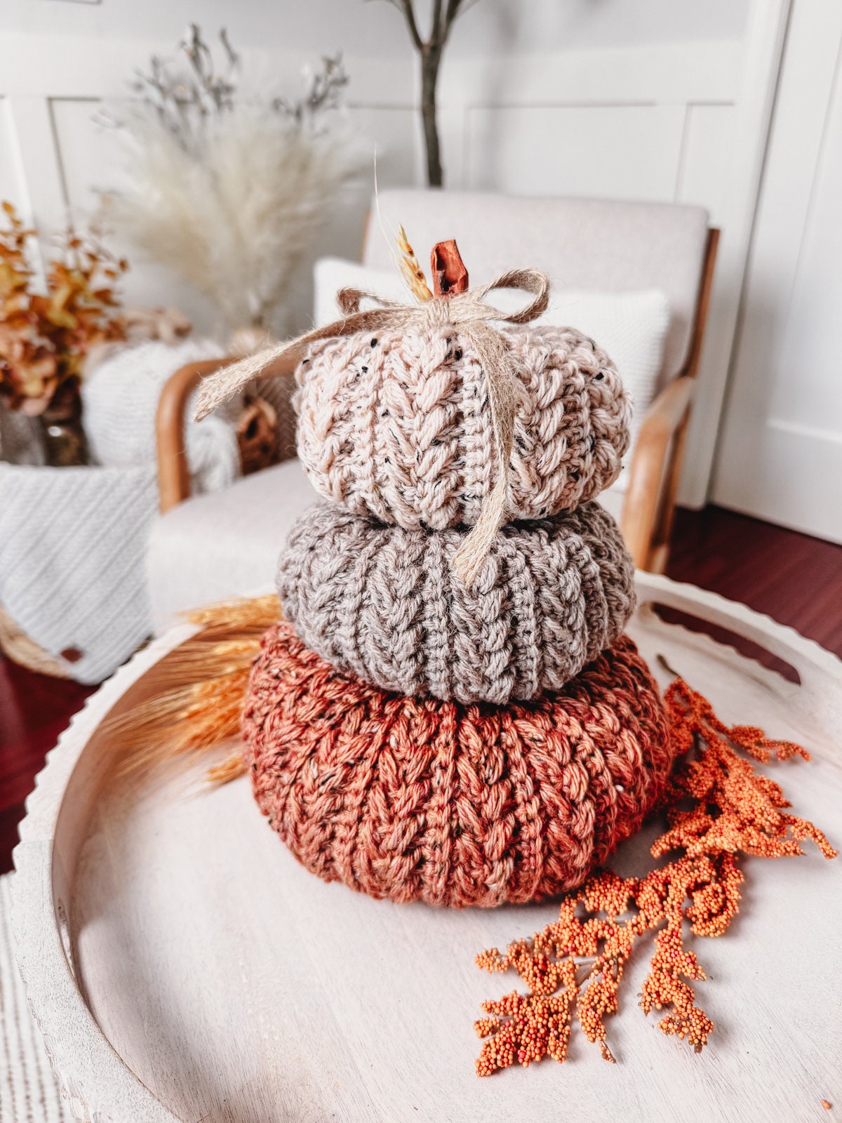 Three knitted pumpkins in varying shades of brown stacked on a white tray, accompanied by orange decorative foliage, make the perfect handmade gift.