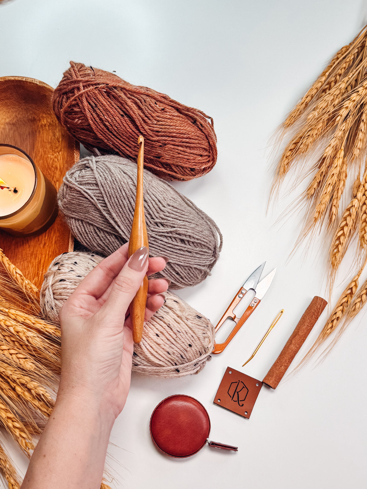 A hand holding a wooden crochet hook with various yarns, a candle, a measuring tape, scissors, and wheat stalks spread on a white surface could easily inspire handmade gifts or unique jewellery creations.
