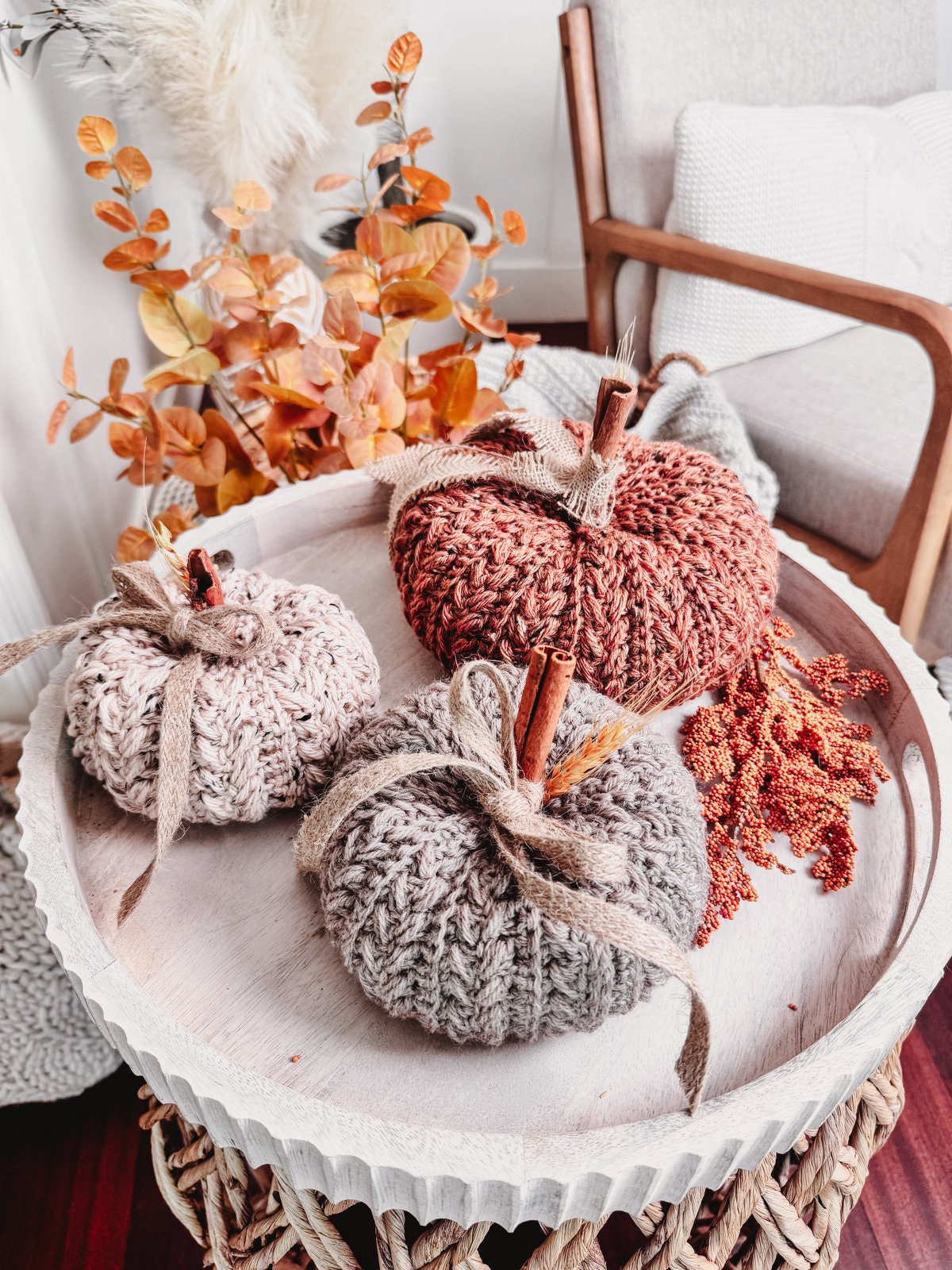 Crocheted pumpkins with cinnamon stick stems are displayed on a round tray, surrounded by autumn foliage, and placed on a woven table—creating the perfect handmade gift.