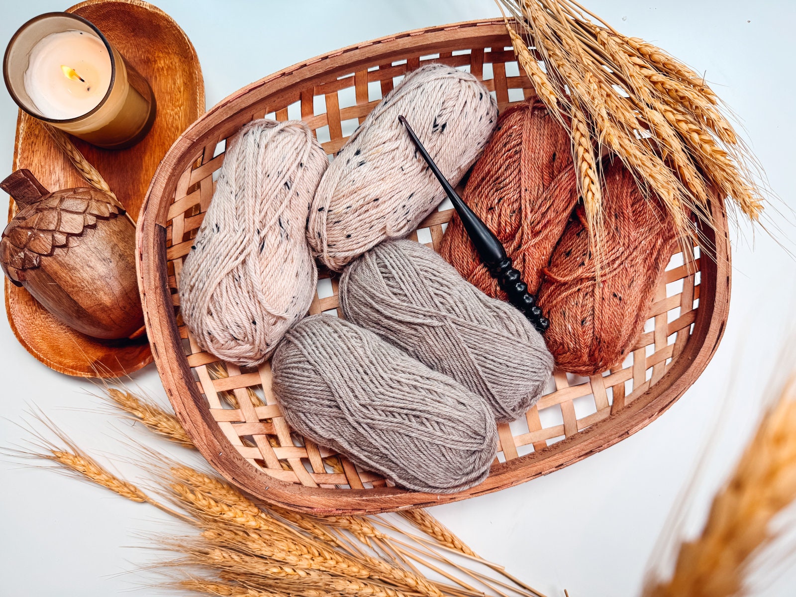 A woven basket holds four skeins of yarn in various shades, a crochet hook, a candle, and wheat stalks on a light surface—perfect for creating a handmade gift.