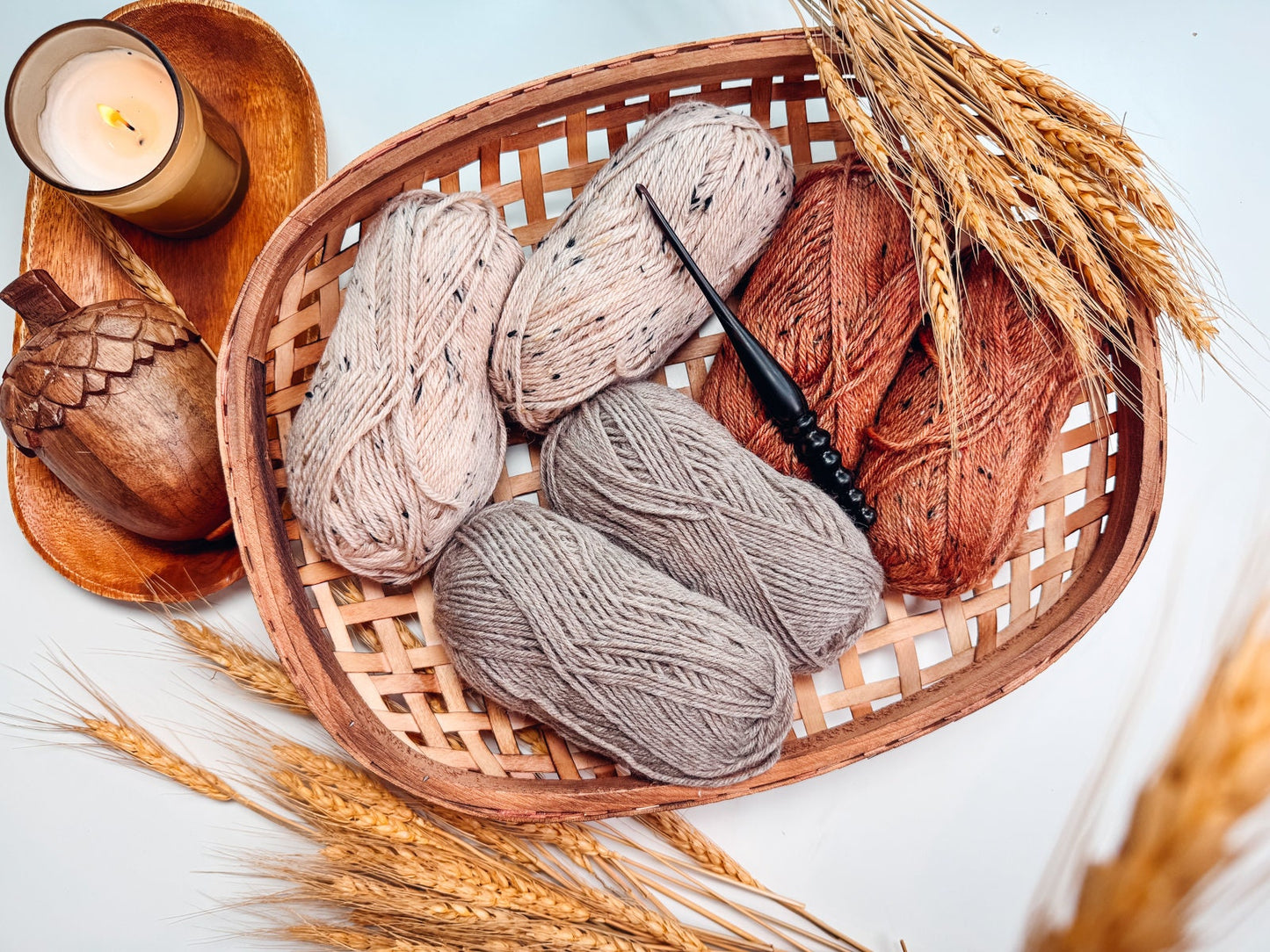 A woven basket holds four skeins of yarn in various shades, a crochet hook, a candle, and wheat stalks on a light surface—perfect for creating a handmade gift.