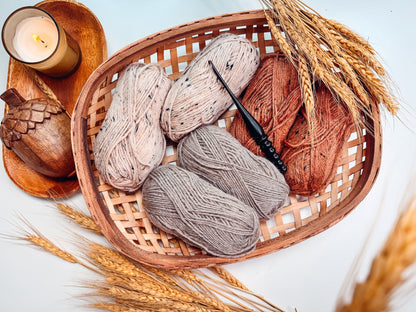 Woven basket holding cream, gray, and rust yarn skeins and a crochet hook, with a candle and wheat stalks nearby