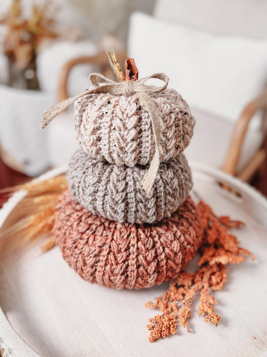 A stack of three knitted pumpkins in shades of beige, gray, and rust is displayed on a wooden tray, adorned with a ribbon and surrounded by dried orange foliage, making it the perfect handmade gift for autumn lovers.