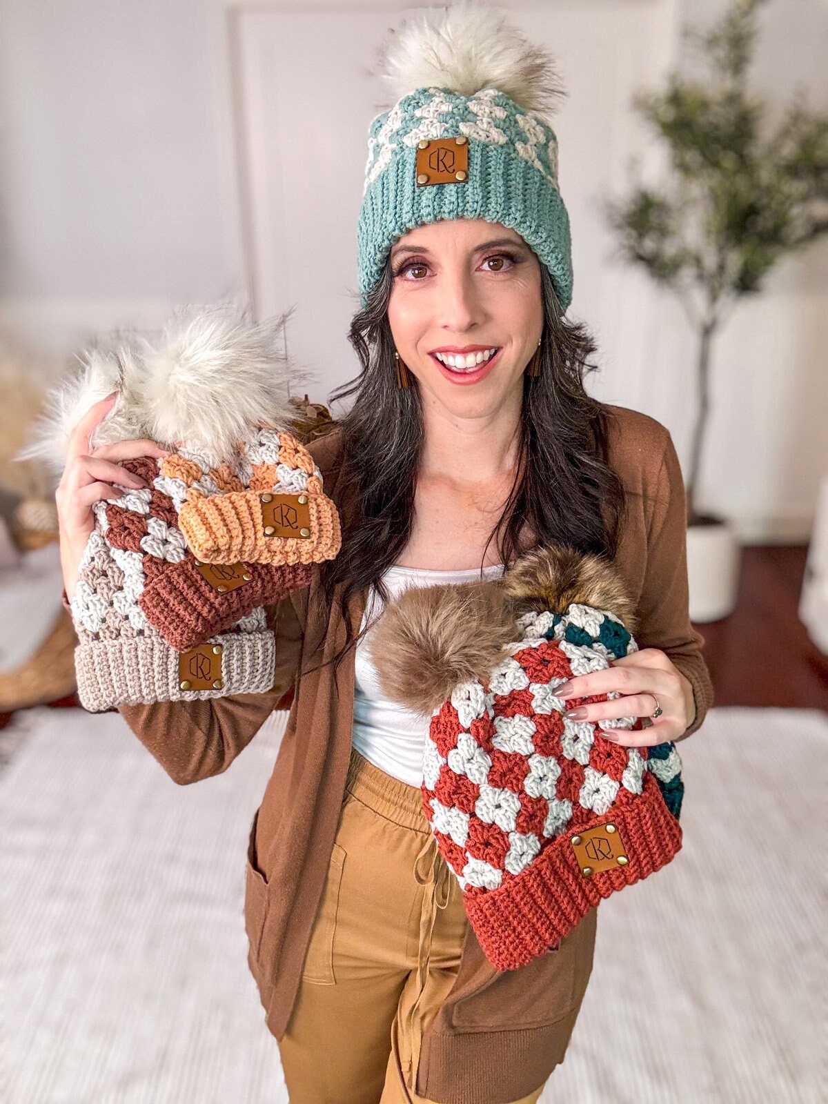 A woman is smiling indoors, holding and wearing multiple knitted hats with pom-poms in a variety of colors. The scene is reminiscent of vintage clothes, with the vibrant hats adding a touch of whimsy to her ensemble.
