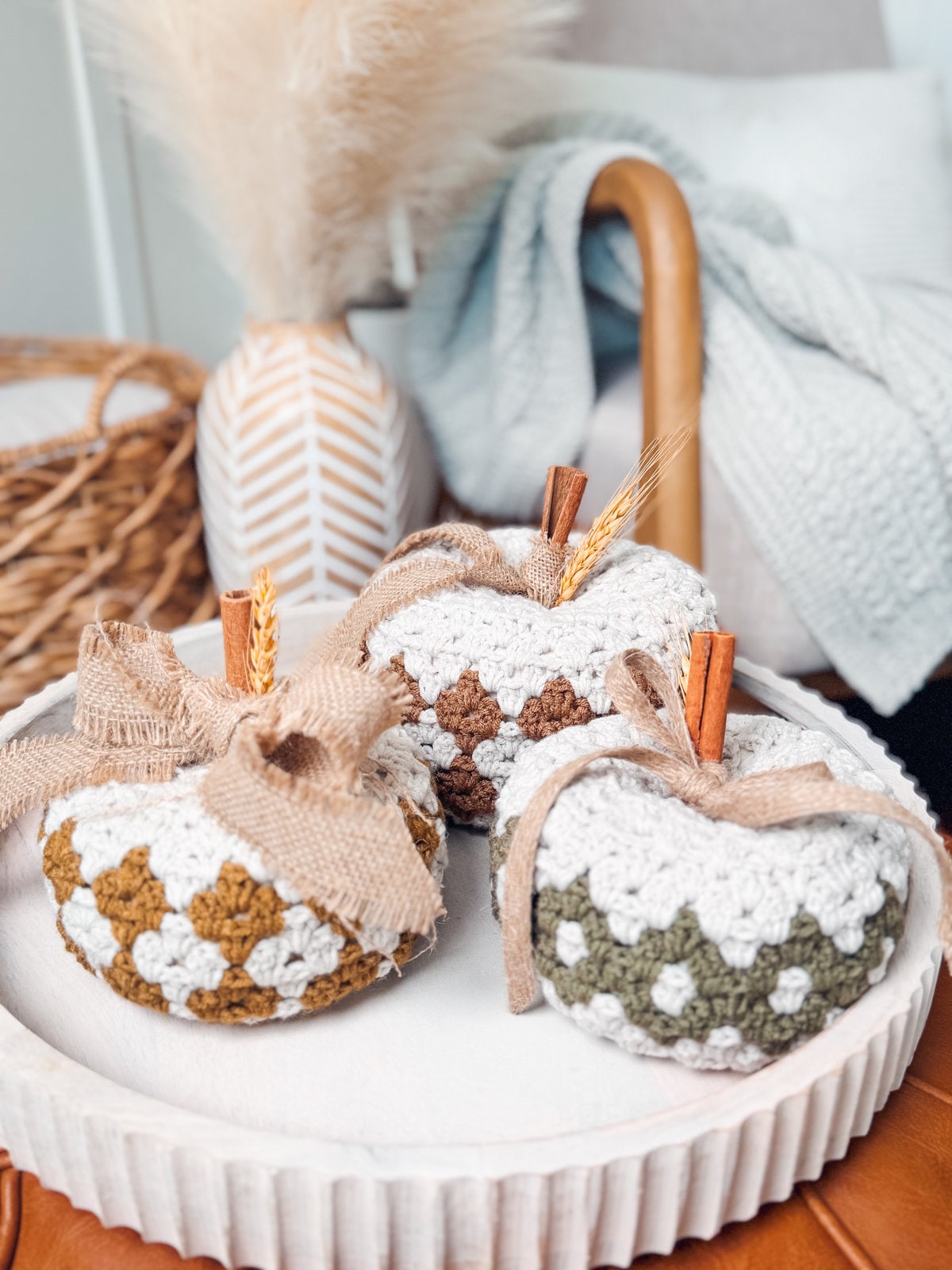 Three crochet pumpkins with a vintage pattern, decorated with cinnamon sticks and burlap ribbons, sit on a white round plate. This handmade gift adds a charming touch of rustic elegance to any setting.