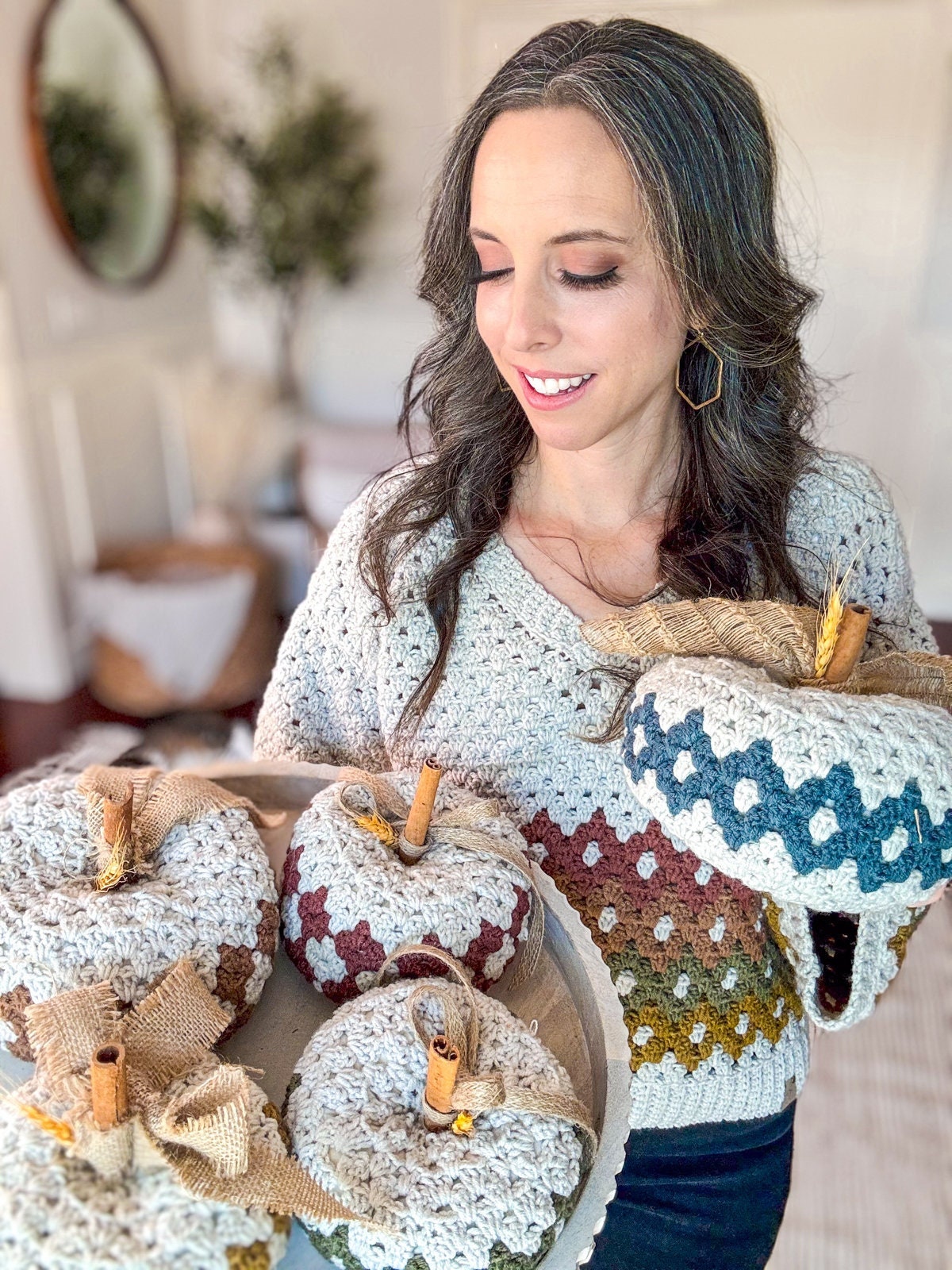 A woman holds a tray of crocheted pumpkins and one in her other hand, all decorated with burlap and cinnamon sticks. She is indoors, wearing a crocheted sweater in matching colors—an ideal setting for presenting a handmade gift.