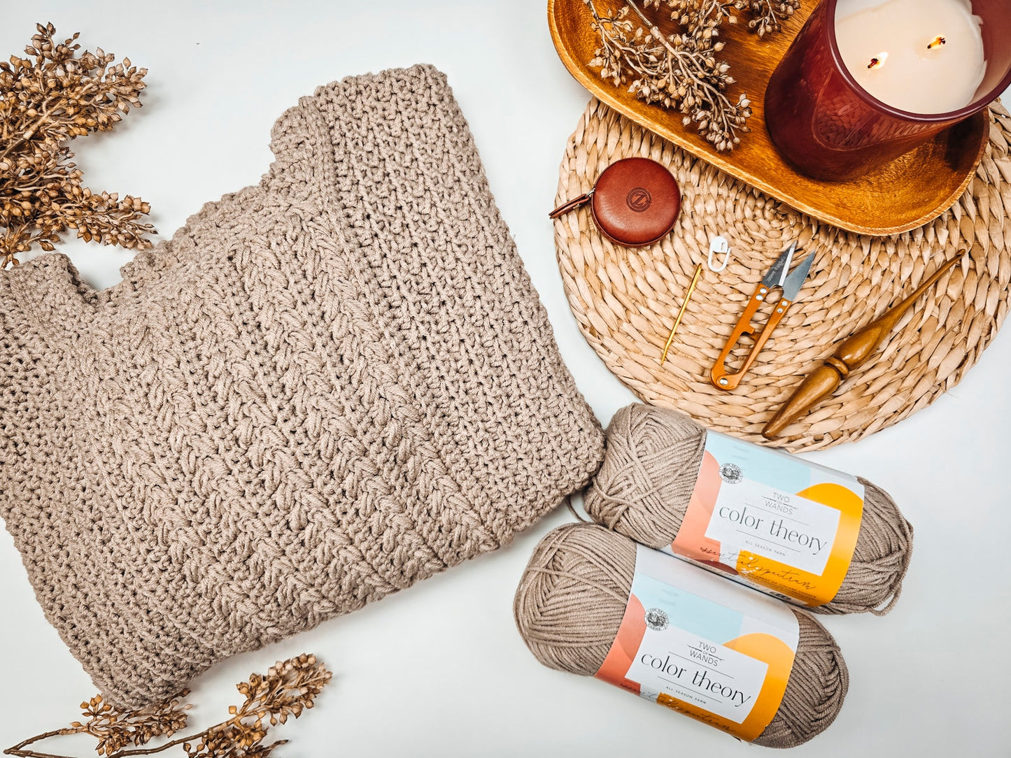 Knitted sweater laid on a white surface surrounded by yarn skeins, knitting tools, a red candle, and dried flowers on a woven mat—a perfect handmade gift reminiscent of vintage clothes.