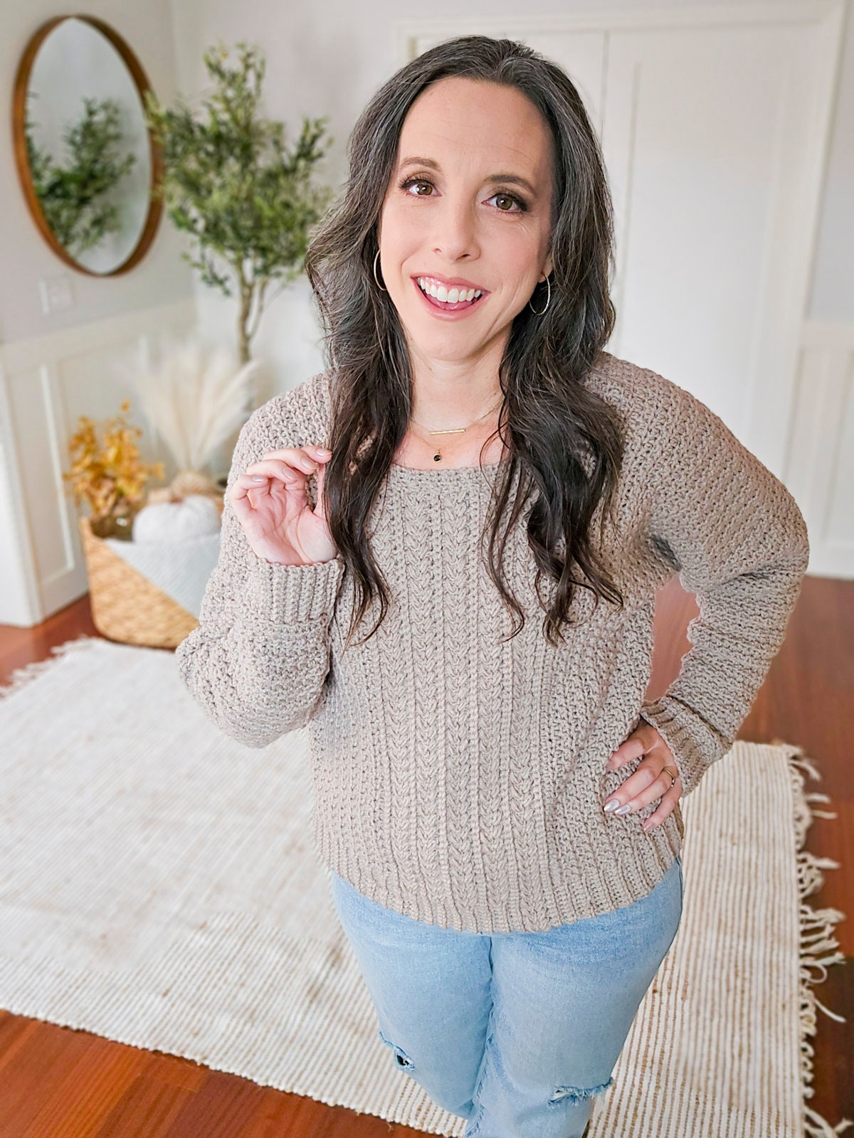 A woman with long dark hair stands in a room with wooden flooring, wearing a beige knit sweater and blue jeans. She showcases unique jewellery that highlights her look. Behind her, a round mirror and a basket with dried plants.