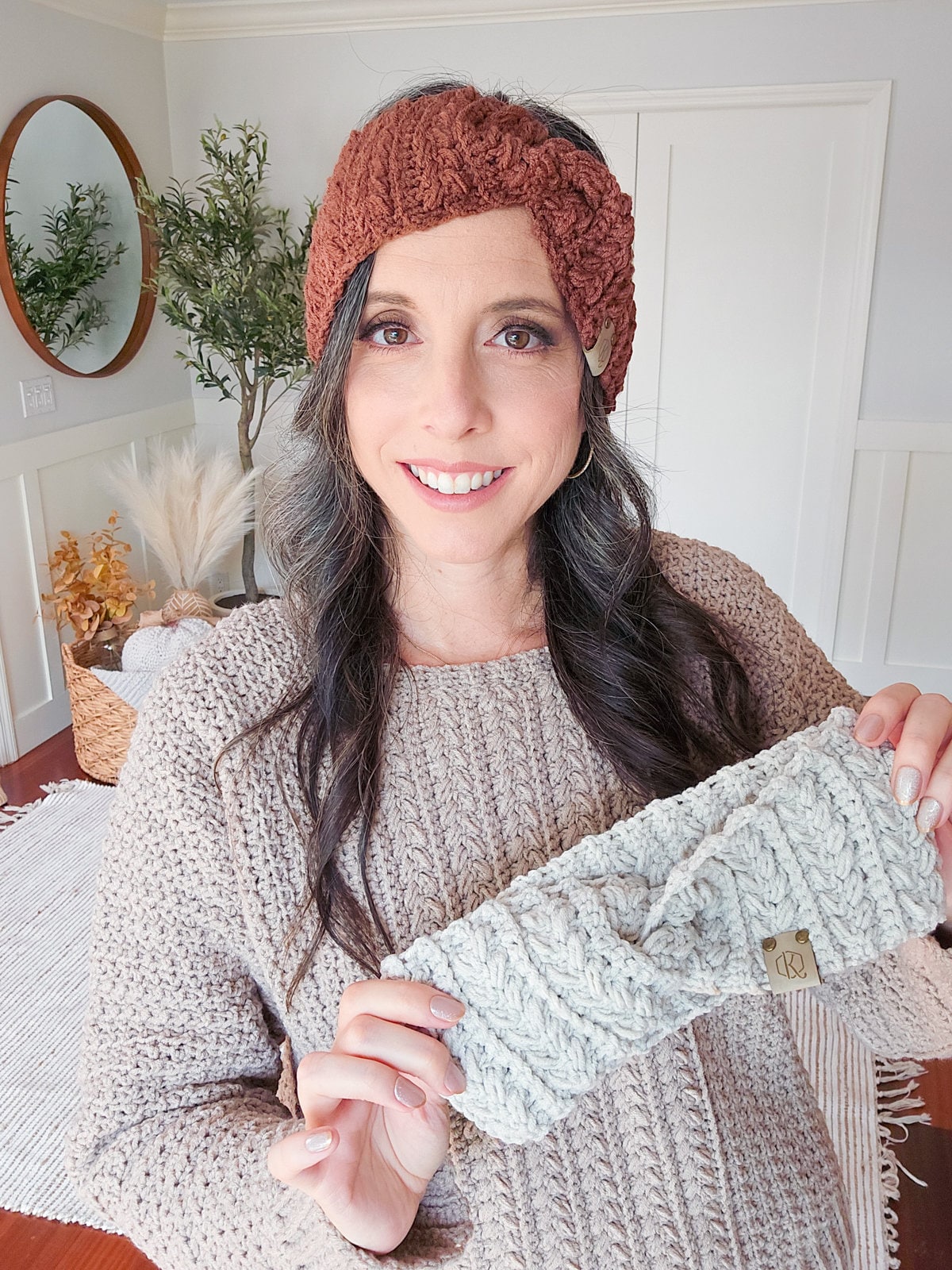 A woman smiling indoors, wearing a knitted ear warmer while holding a second handmade gift ear warmer in her hand.