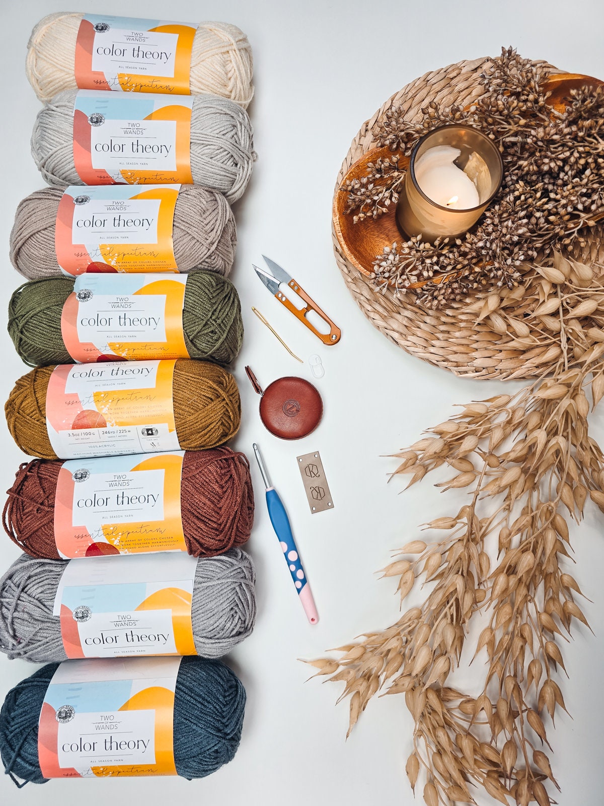 A selection of colorful yarns arranged vertically, alongside knitting tools, a candle in a woven holder, and dried foliage on a white surface, creating the perfect backdrop for unique jewellery or handmade gifts.