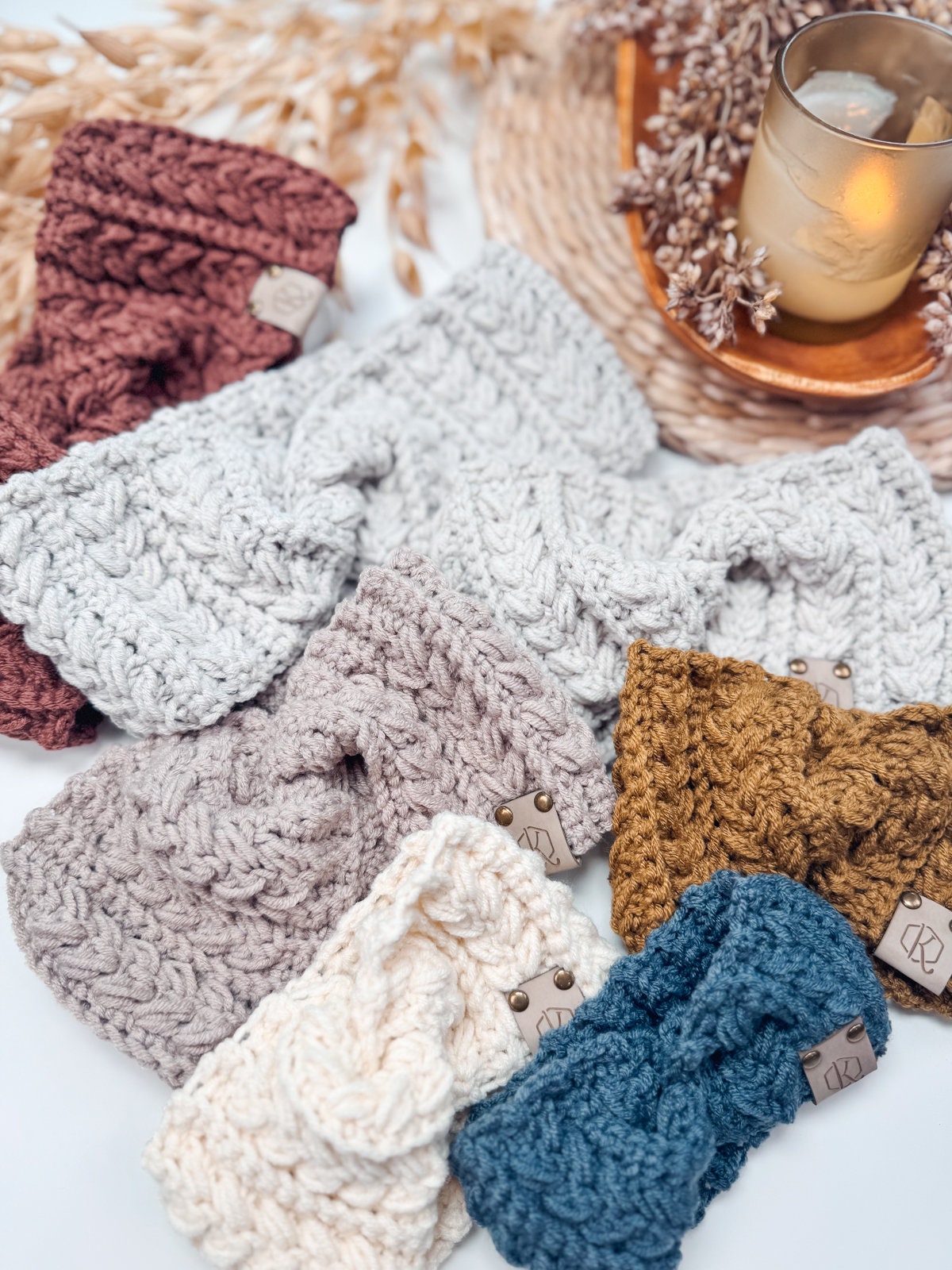 A variety of knitted headbands in different colors are arranged next to a lit candle and decorative dried plants, making it an ideal setting for showcasing unique jewellery or selecting a handmade gift.
