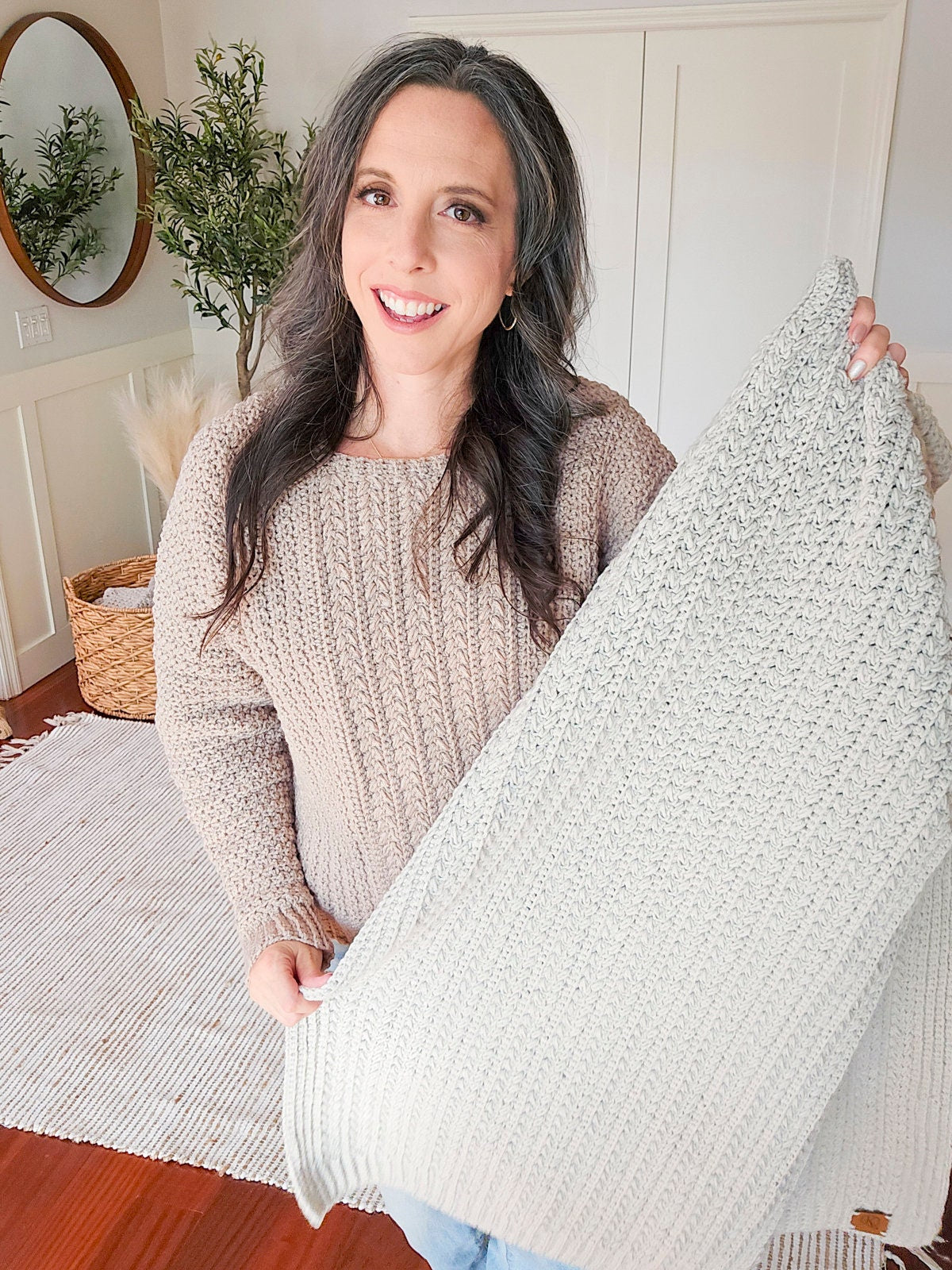 A person in a beige sweater holds a light-colored woven blanket, smiling in a room with a plant, circular mirror, and unique jewellery displayed on the table.