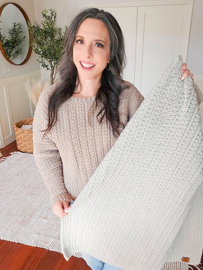 Smiling woman in a tan sweater holds up a light gray crochet blanket with braided texture in a bright room