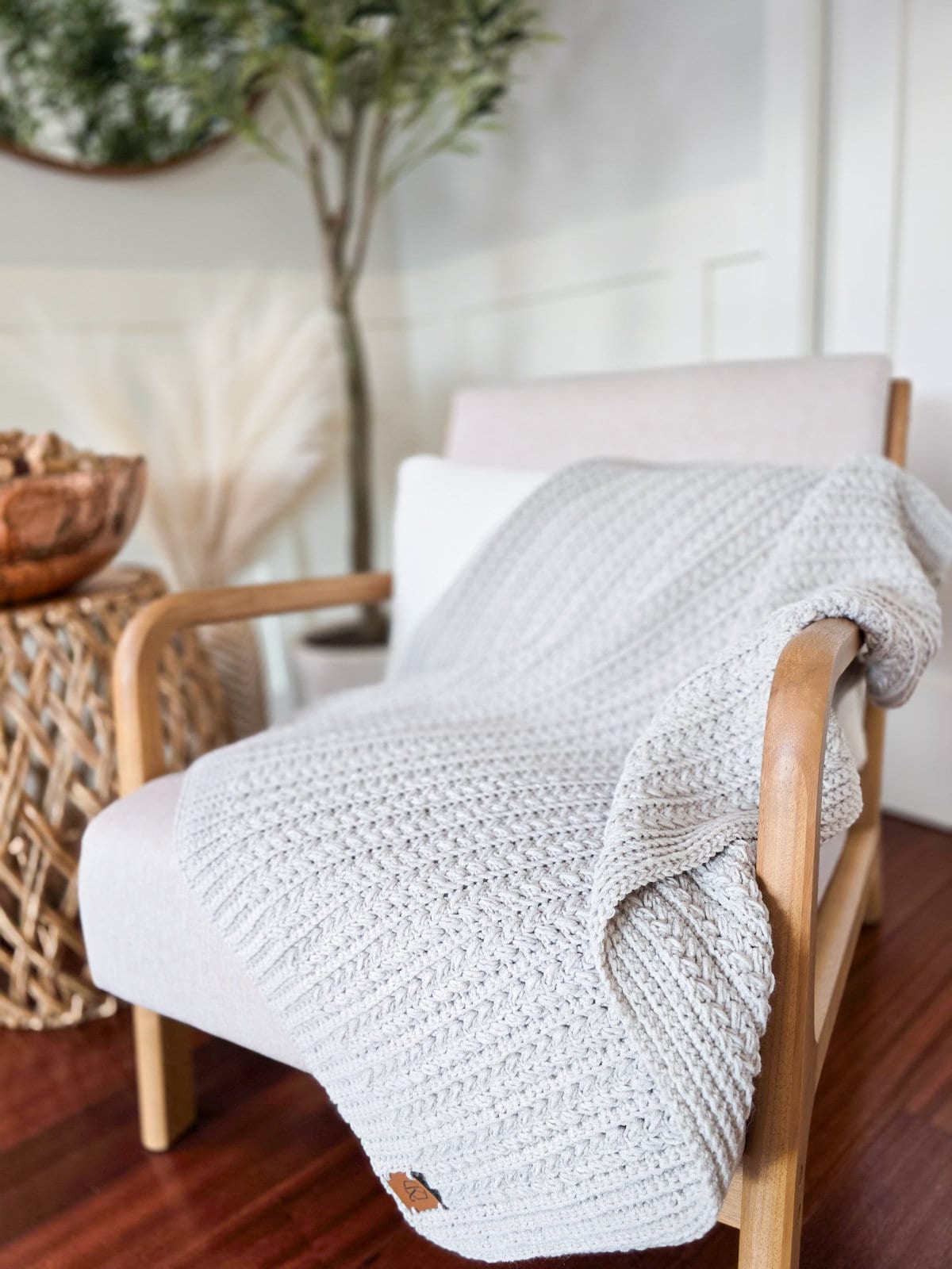 A white armchair with light wooden arms, draped with a textured grey crochet blanket, sits beside a wicker side table adorned with a handmade gift. In the background, a bowl of pinecones and potted plant complete the cozy scene.