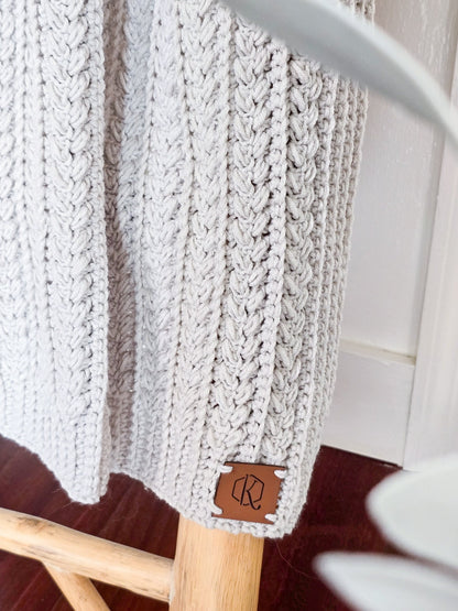 Close-up of light gray crochet blanket with braided columns and a leather logo tag on a wooden ladder