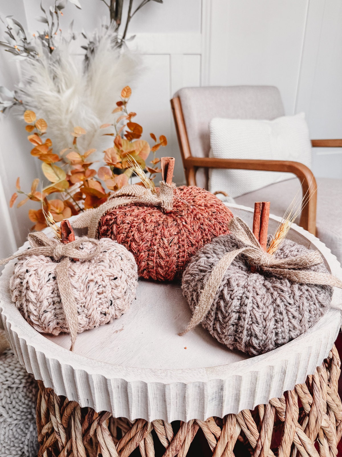 Three knitted pumpkins in shades of beige, burnt orange, and gray sit on a round white tray, complemented by autumnal foliage and a cozy chair in the background.