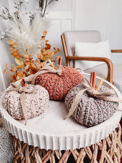 Three crochet pumpkins in cream, rust, and gray with cinnamon stick stems on a white tray with fall leaves