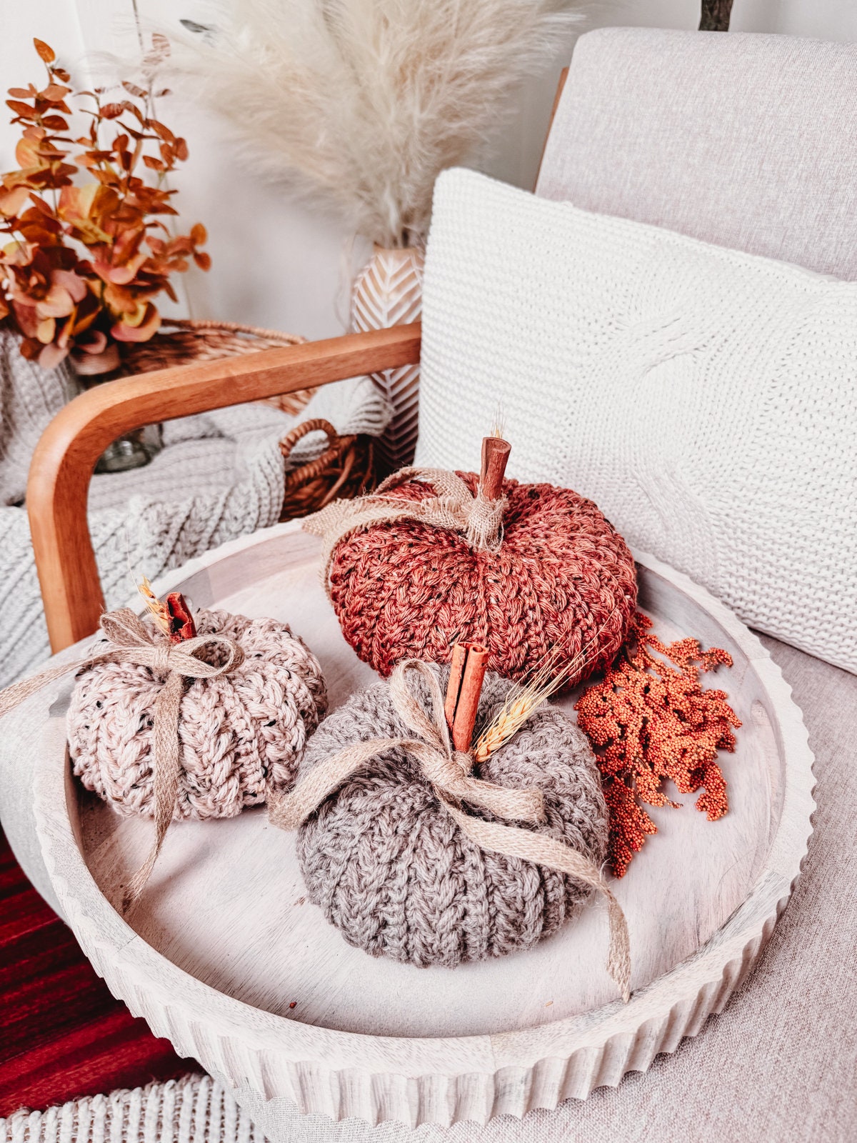 A wooden tray on a chair holds three knitted pumpkins in varying sizes and colors, adorned with cinnamon sticks and twine. Autumnal decor, including a bouquet of dried leaves and unique jewellery, adds a touch of charm to the background.