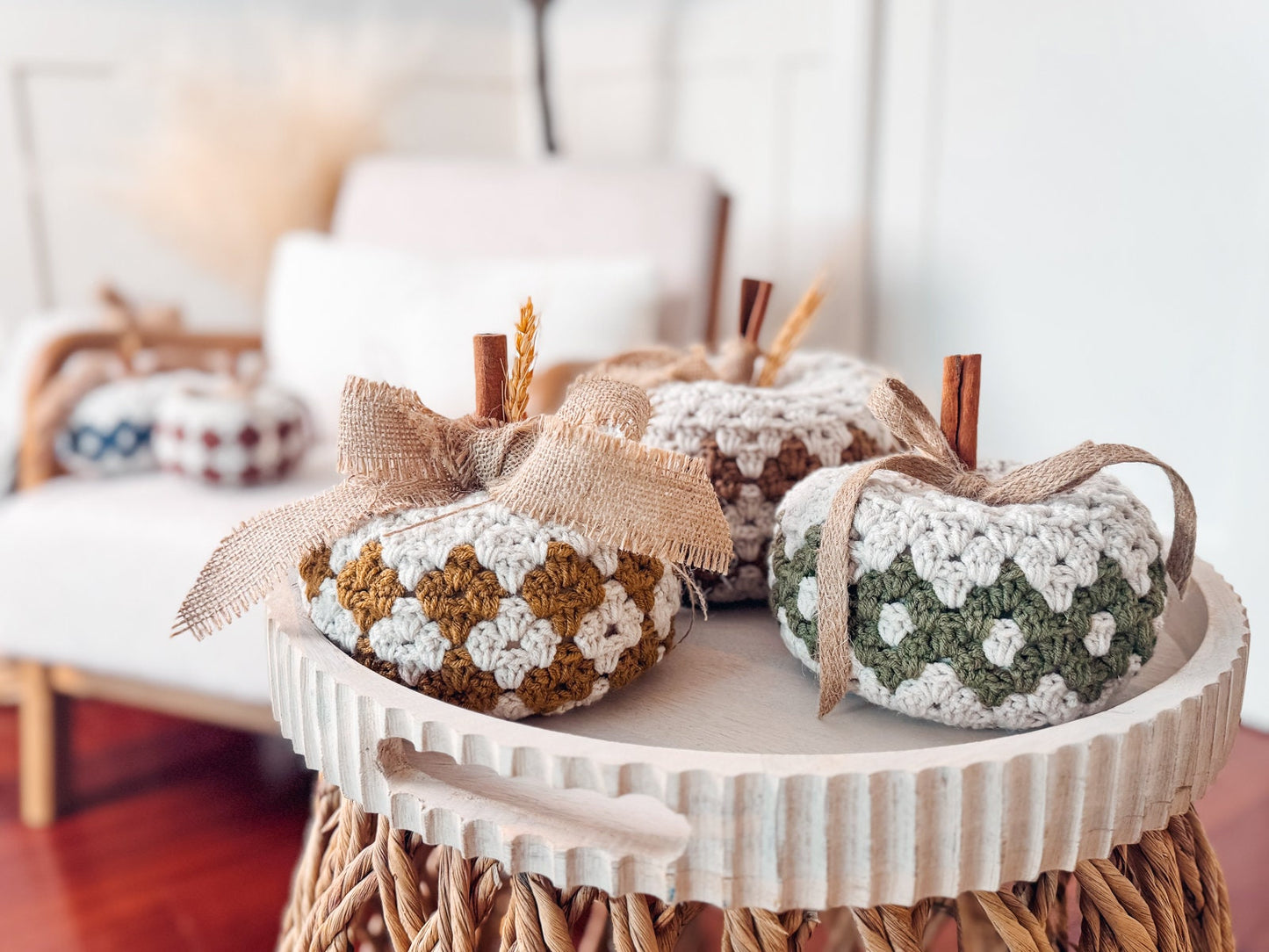 Three crocheted pumpkins with ribbon bows sit on a round white table, perfect as a handmade gift, in front of a cozy chair and yarn basket in the background.