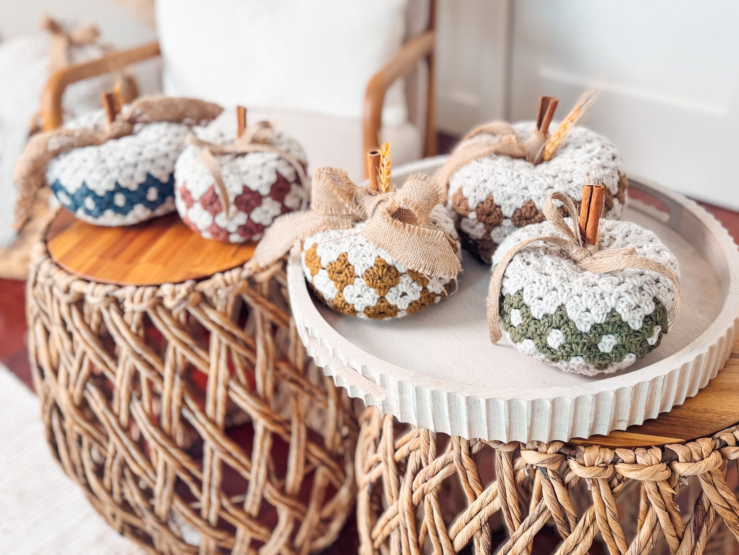 A display of five crocheted pumpkins with cinnamon stick stems, sitting on woven and wooden tables. Each pumpkin is decorated with different colored patterns and burlap bows, making a charming handmade gift.