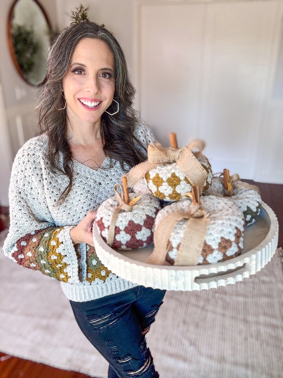 A woman with long hair and a sweater holds a round tray of crocheted pumpkins, perfect as a charming handmade gift.