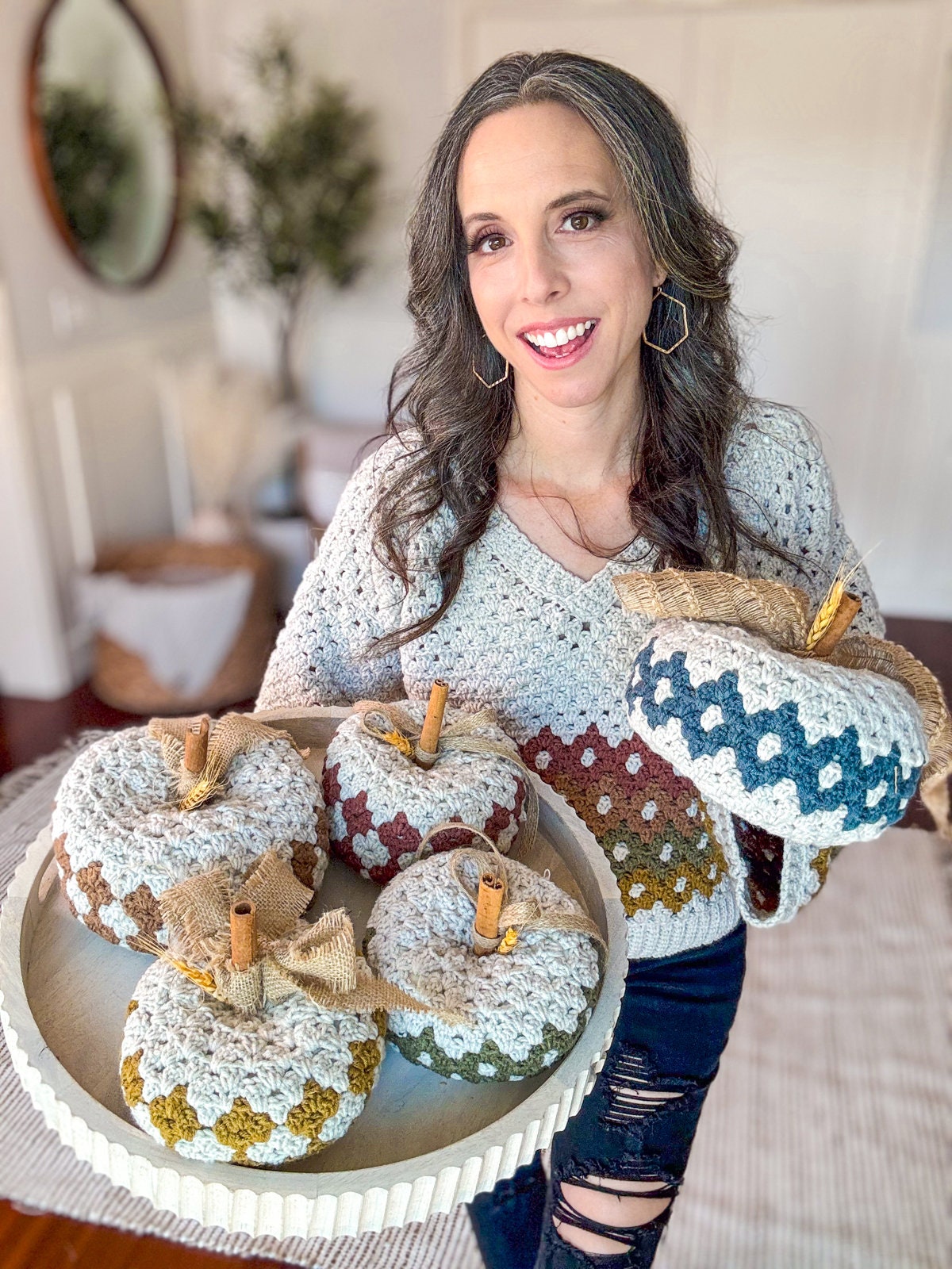A person wearing a crocheted sweater holds a tray with crocheted pumpkins in various colors, accompanied by pieces of unique jewellery.