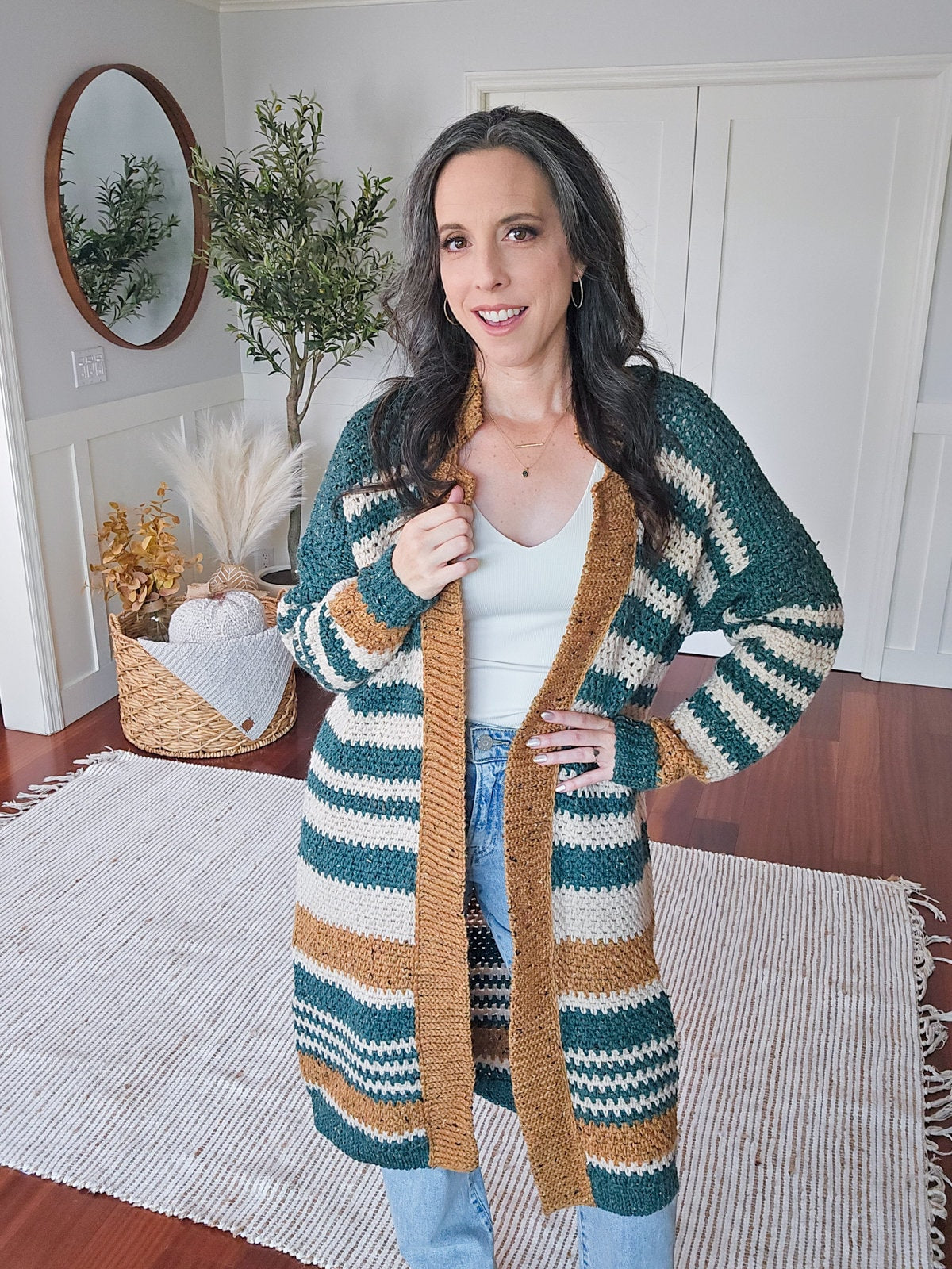 A person stands on a rug in a living room, wearing a striped cardigan, white shirt, and jeans. A mirror, plant, and woven basket in the background. The outfit is accentuated by unique jewellery that adds an extra touch of individuality to the scene.