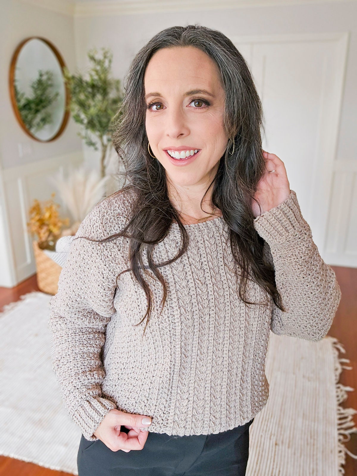 A woman with long hair and a gray knit sweater smiles at the camera in a well-lit room with a round mirror, plants, and unique jewellery displayed in the background.