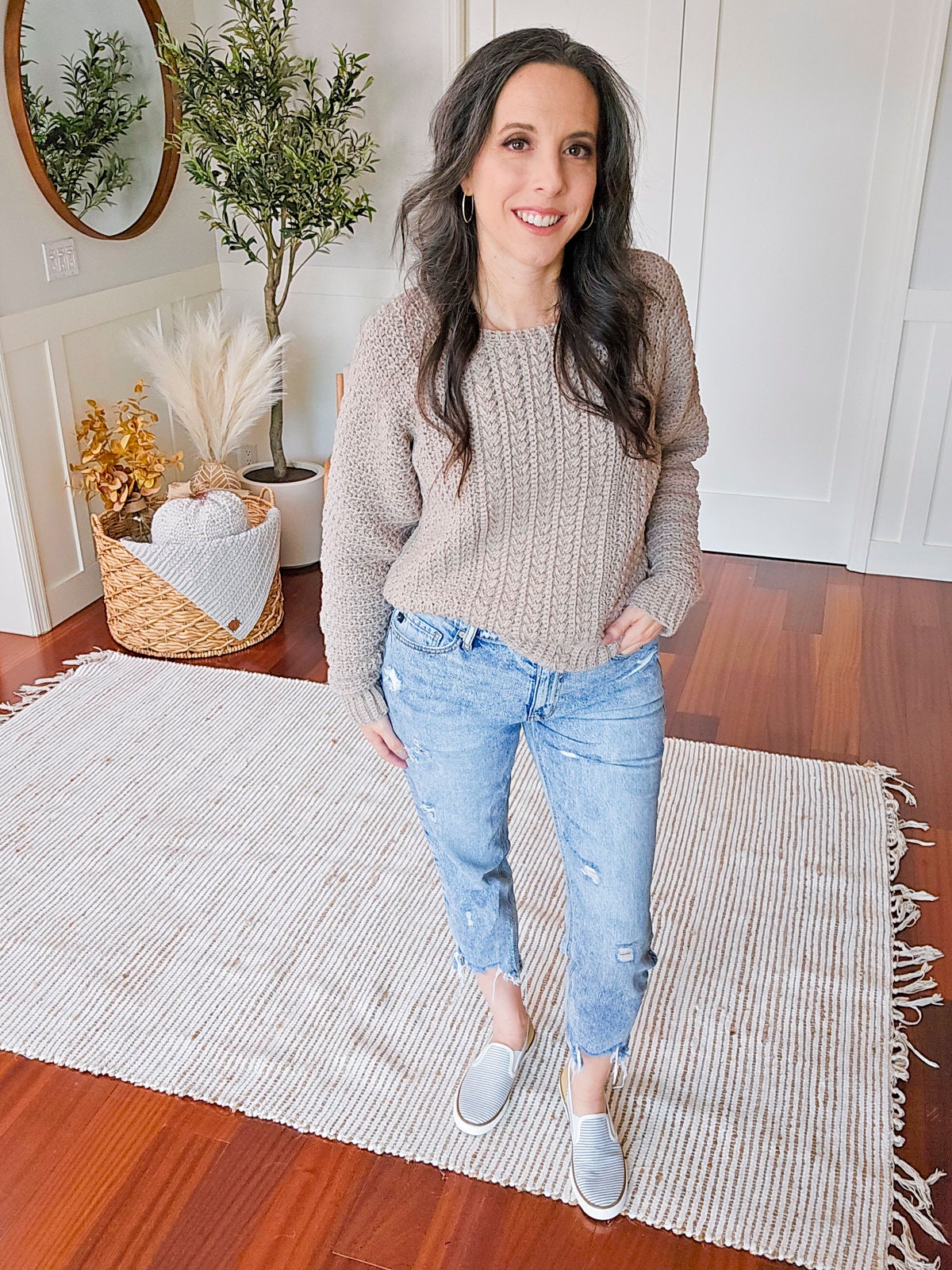 A woman stands on a rug in a room with wooden flooring, wearing a beige knit sweater, blue jeans, and grey slip-on shoes. She complements her look with unique jewellery. In the background, there is a plant and a decorative basket.