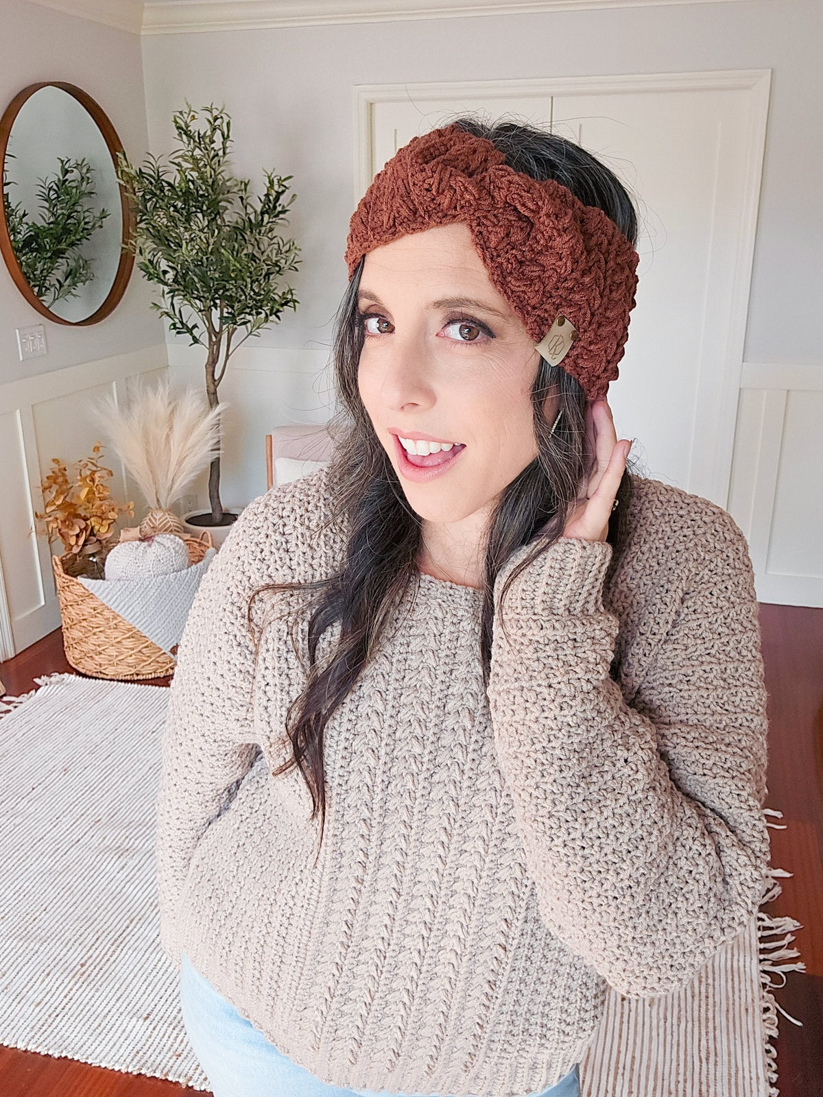 Person wearing a knitted headband and a textured sweater, posing indoors with unique jewellery. A basket of dried plants and a round mirror are in the background.
