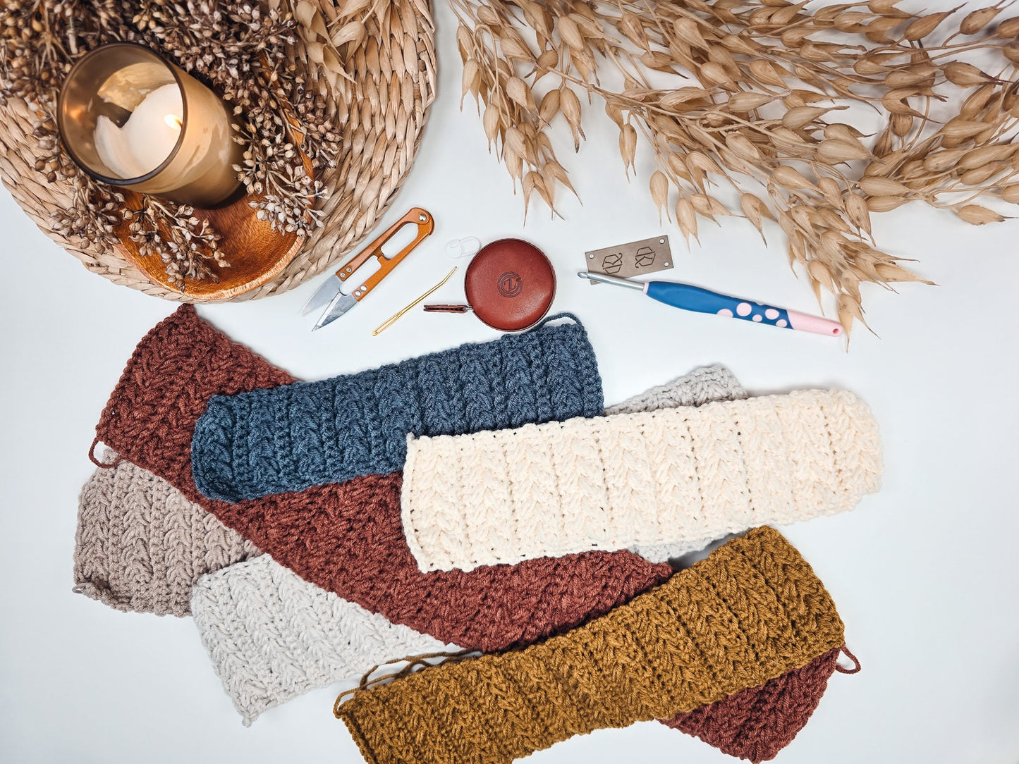 A display of crocheted fabric pieces in various colors, placed on a white surface with crochet tools, a lit candle, and dried plants—making for the perfect handmade gift.