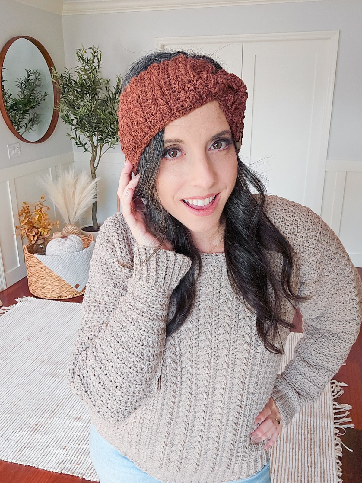 A woman with long dark hair wearing a knit sweater, a brown knit headband, and vintage clothes smiles, standing indoors in a room with a round mirror, a plant, and a basket of dried flowers.